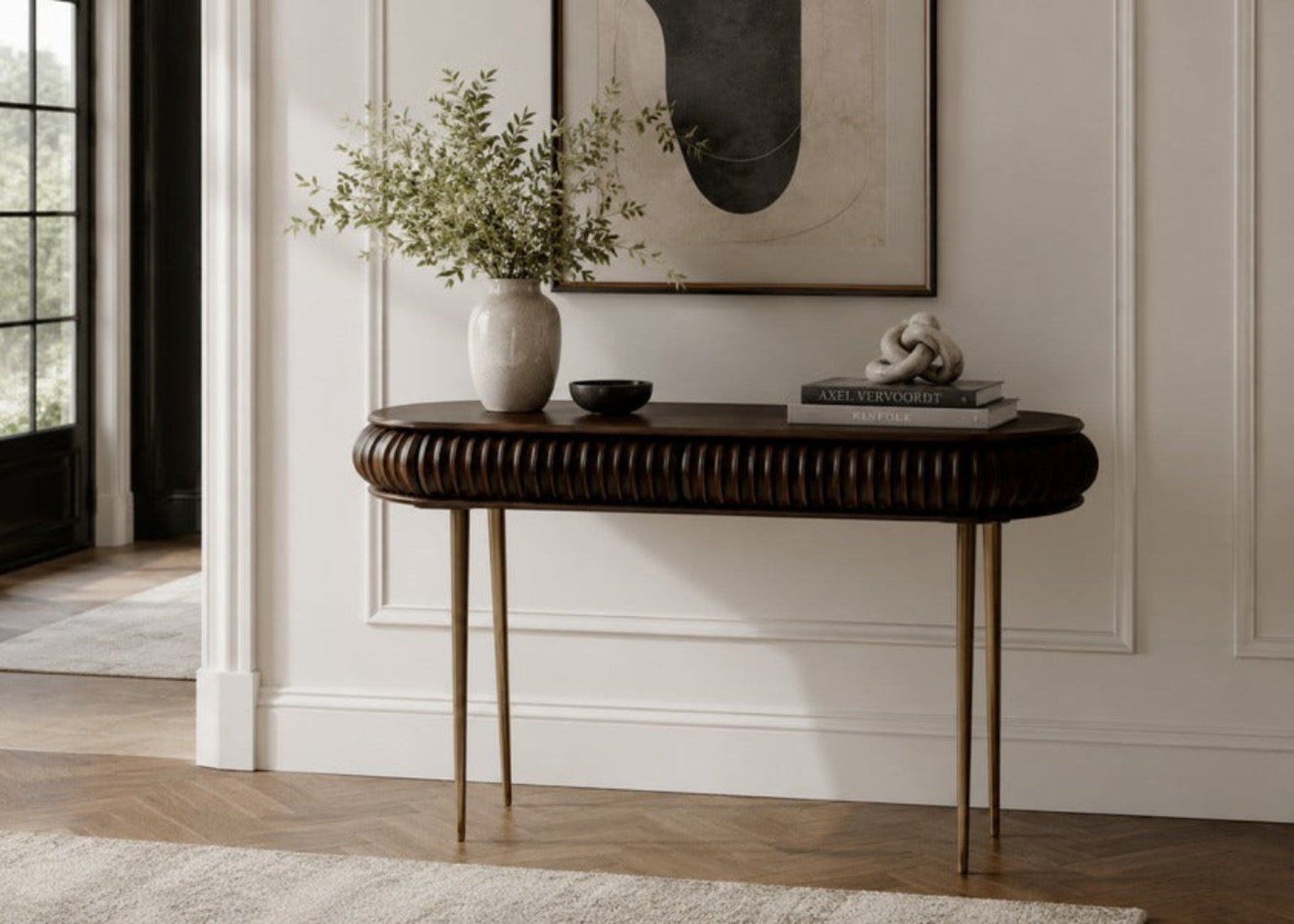 A dark wood console table with a ribbed oval top and slender gold-toned legs, styled in a bright entryway with a white vase of greenery, a small bowl, and a decorative sculpture on top.